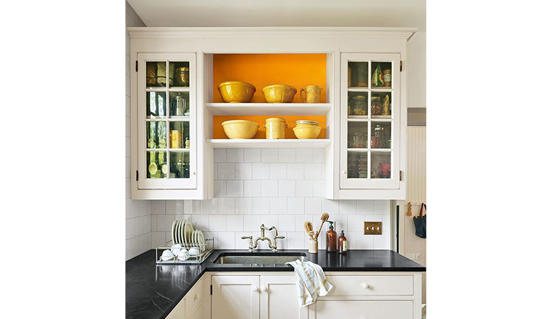 A mostly white-painted kitchen with black counter tops and orange accent behind shelving. A mostly white-painted kitchen with black counter tops and orange accent behind shelving.